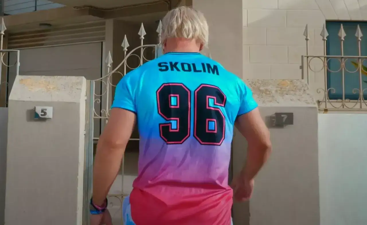 Mężczyzna w koszulce z napisem SKOLIM i numerem 96, stojący przed domem z numerem 5.