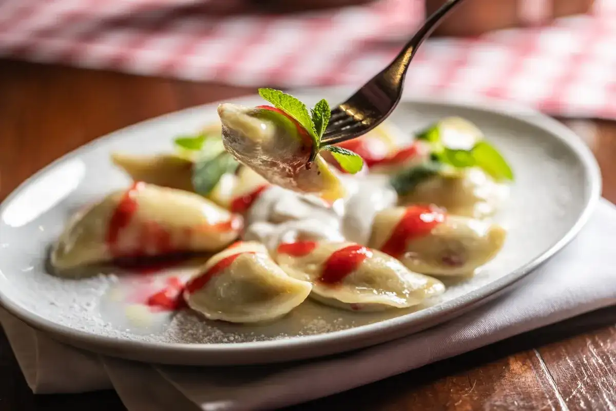 Jak gotować pierogi mrożone? Na talerzu pyszne pierogi z polewą truskawkową i miętą, jeden pieróg na widelcu.