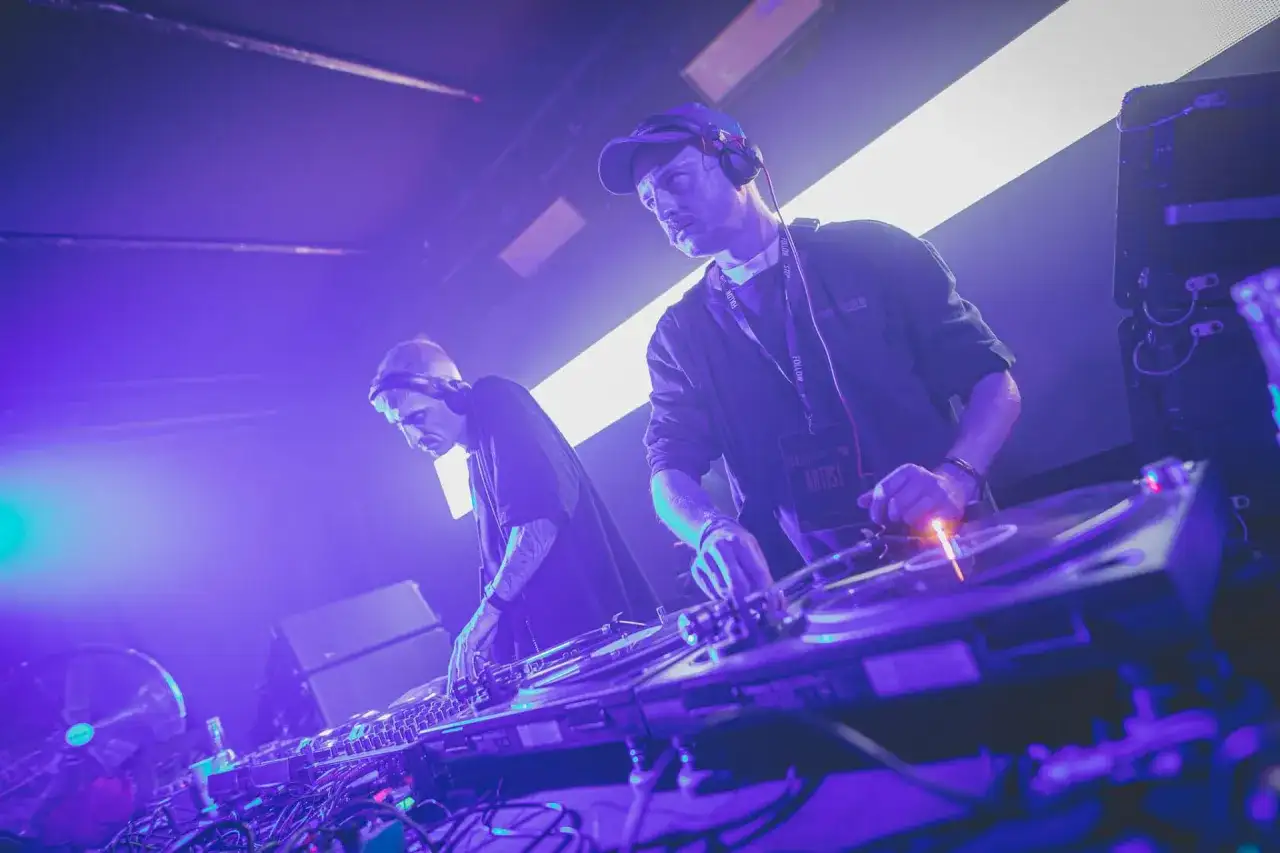Dwóch DJ-ów w słuchawkach, tworzących muzykę techno. Jeden z nich ma na szyi identyfikator "ARTIST".