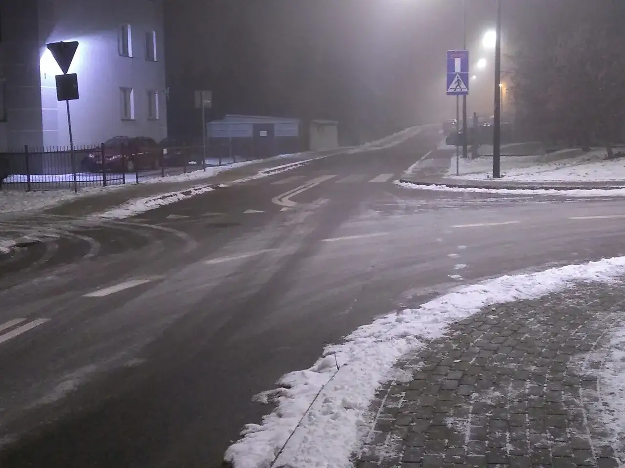 Skrzyżowanie dróg w nocy, z mgłą, śniegiem i znakami drogowymi.
