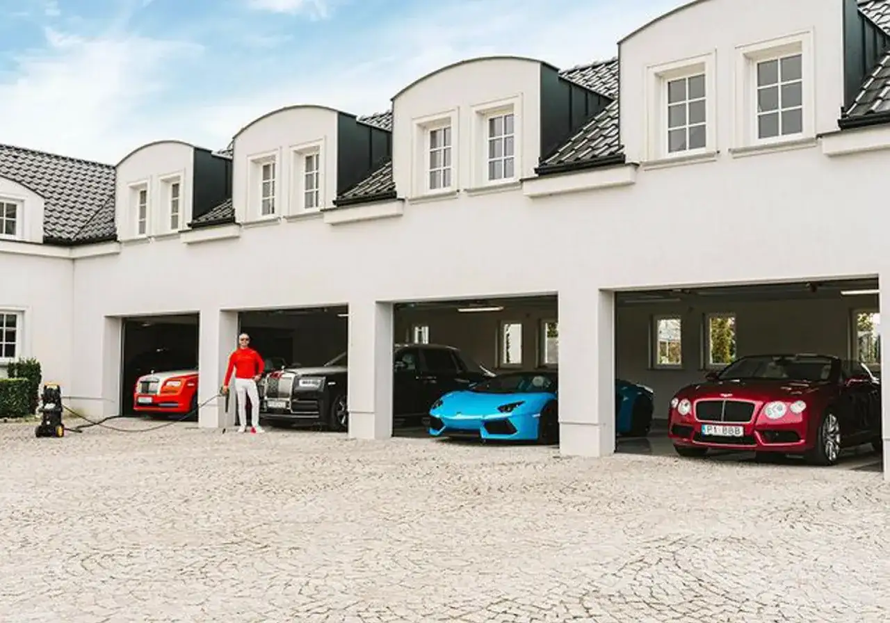 Piotr Śledź w czerwonej bluzie i białych spodniach stoi przy luksusowych samochodach: czerwonym Rolls-Royce, czarnym Rolls-Royce, niebieskim Lamborghini i czerwonym Bentleyu.