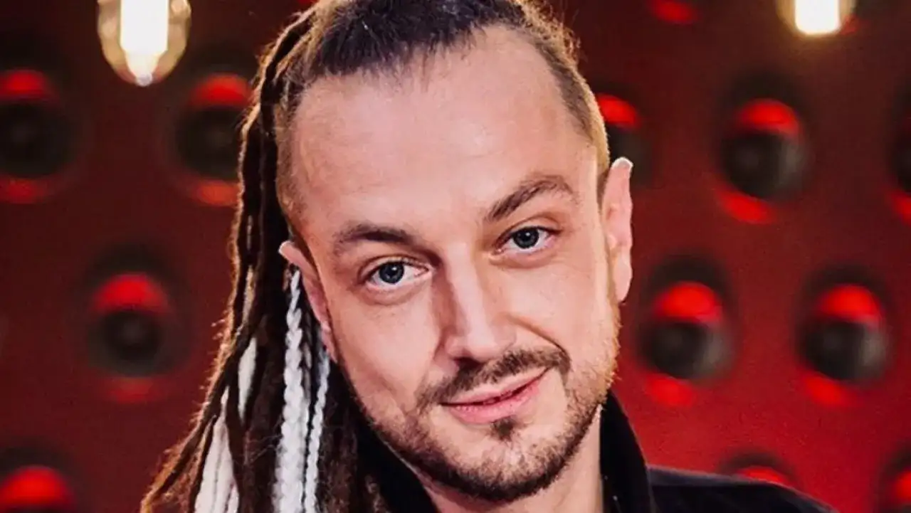 Aleksander Milwiw Baron, w wieku około 40 lat, z dredami i niebieskimi oczami, uśmiecha się na tle czerwonej ściany.