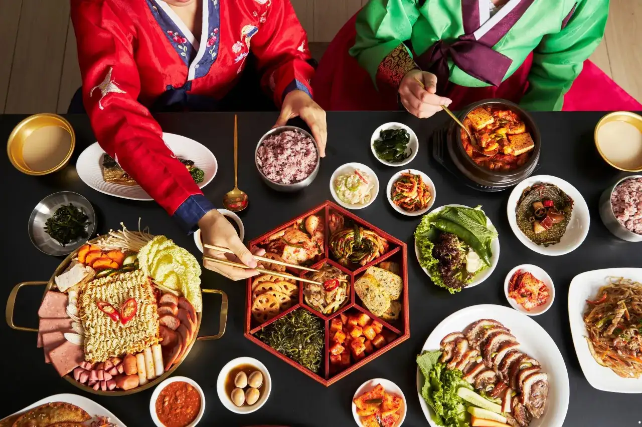 Dwie osoby w tradycyjnych koreańskich strojach jedzą obfity posiłek z wieloma daniami, w tym kimchi.