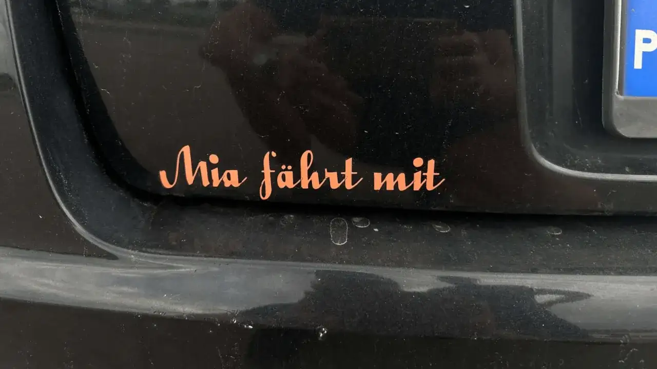 Pomarańczowa naklejka na czarnym lakierze samochodu z napisem "Mia fährt mit". Chcesz wiedzieć, jak usunąć naklejki z samochodu?