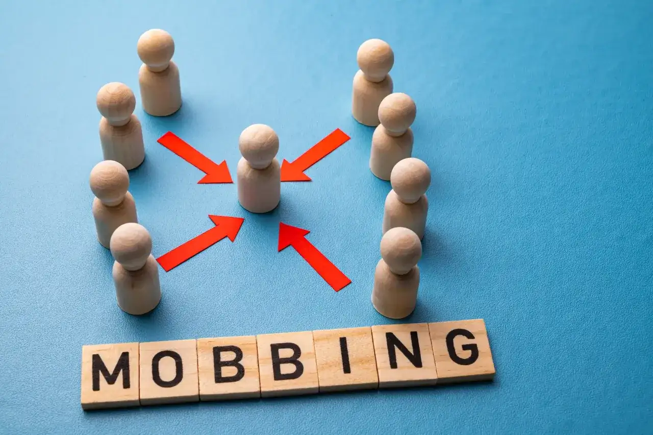 Drewniane ludziki otaczają jednego, a czerwone strzałki wskazują na niego. Słowo "MOBBING" na klockach.