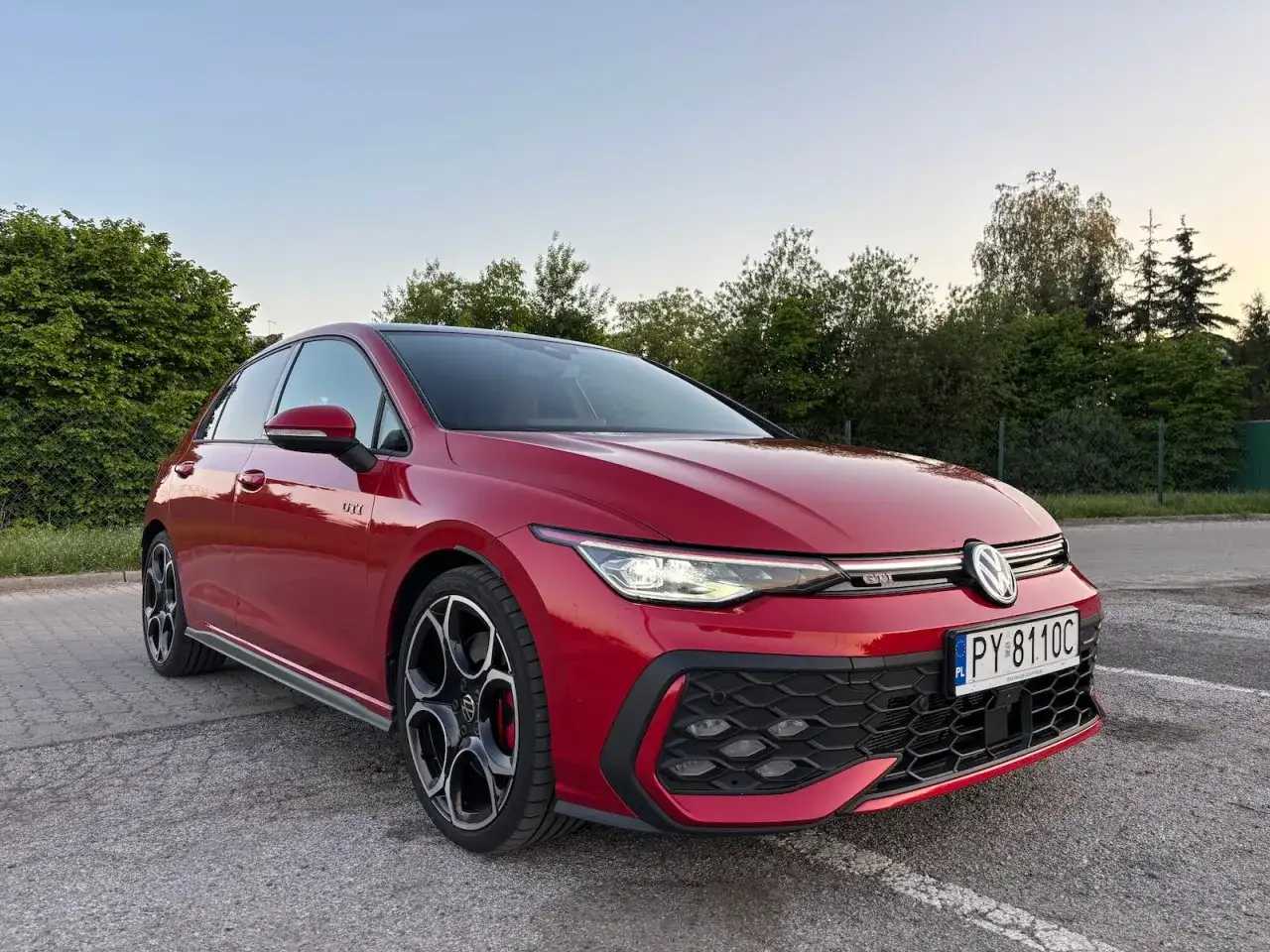 Czerwony Volkswagen Golf GTI na parkingu, z zielenią w tle.