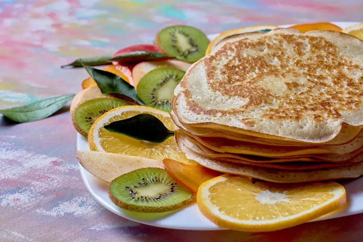 Stos naleśników z budyniem, otoczony plasterkami kiwi, pomarańczy i jabłka, ozdobiony listkami.