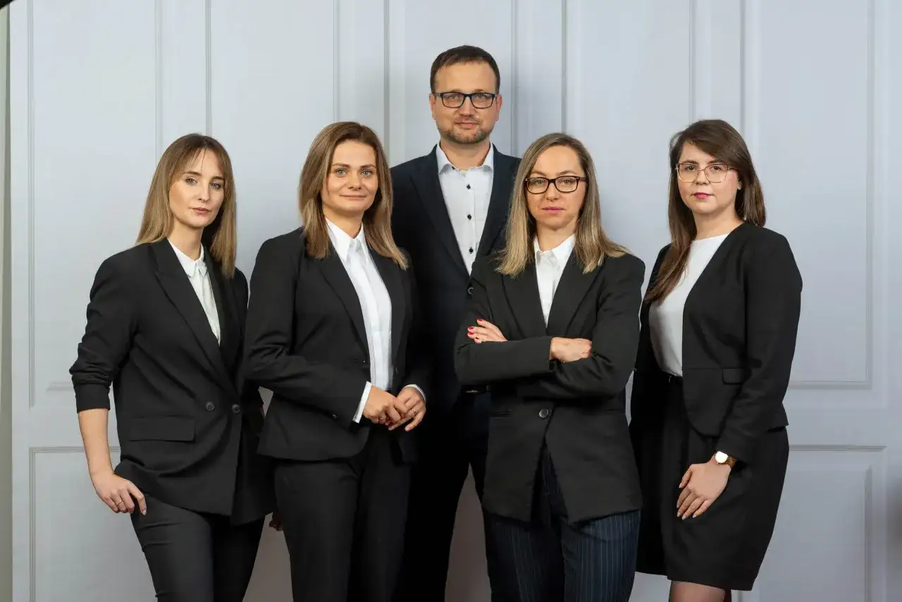 Pięć osób w garniturach, zespół prawników, stoi przed jasną ścianą.
