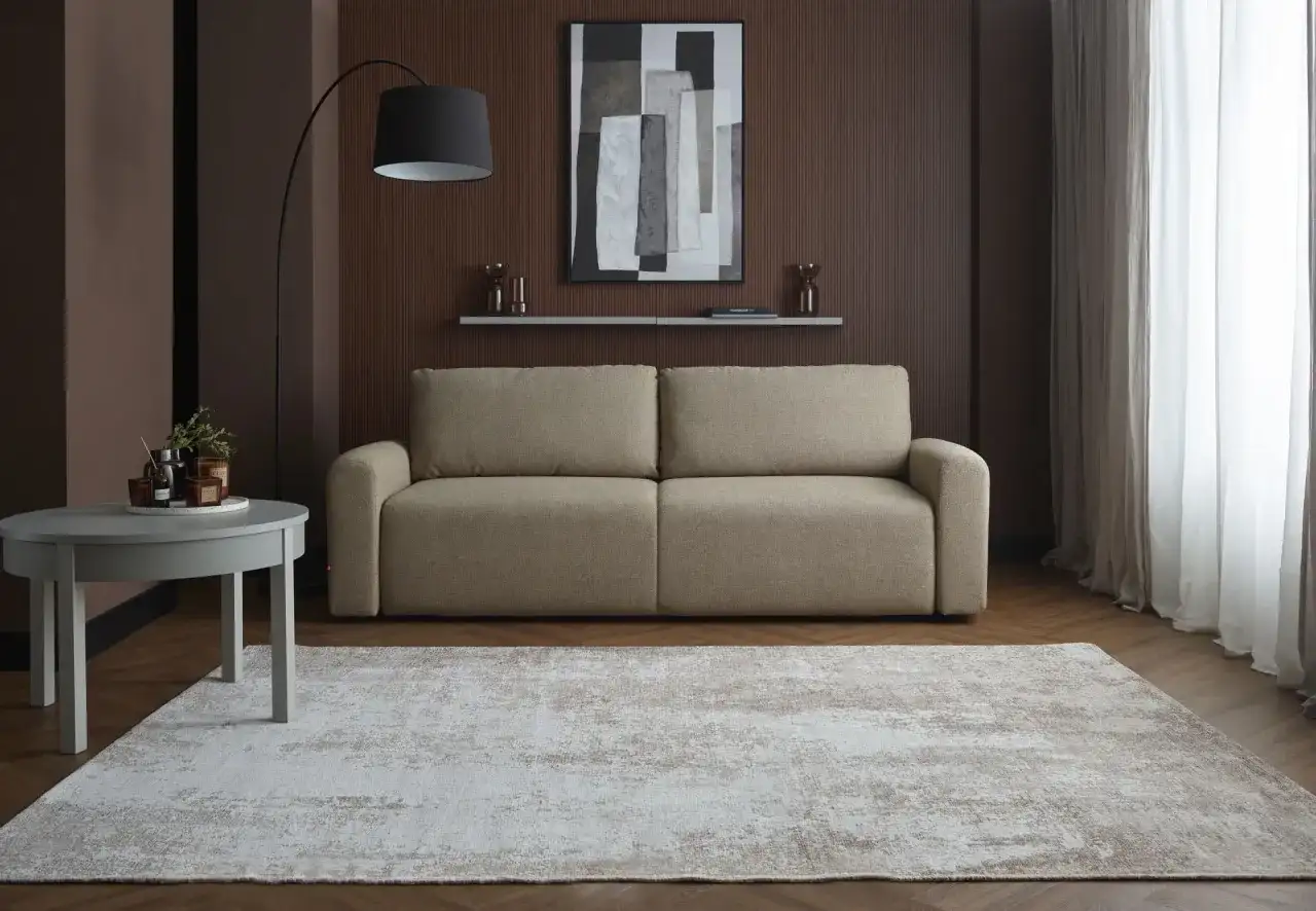 Elegancka, beżowa kanapa na tle brązowej, panelowej ściany w salonie. Nad nią abstrakcyjny obraz i półka z dekoracjami.