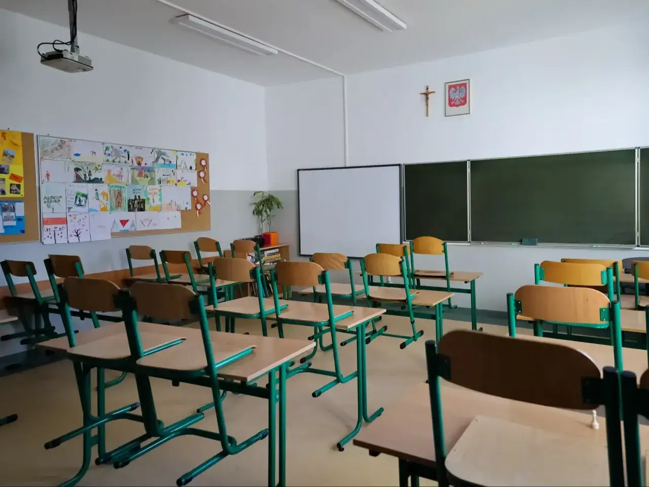 Pusta sala lekcyjna z rzędami krzeseł i biurek, tablicą i dziecięcymi rysunkami na ścianie.