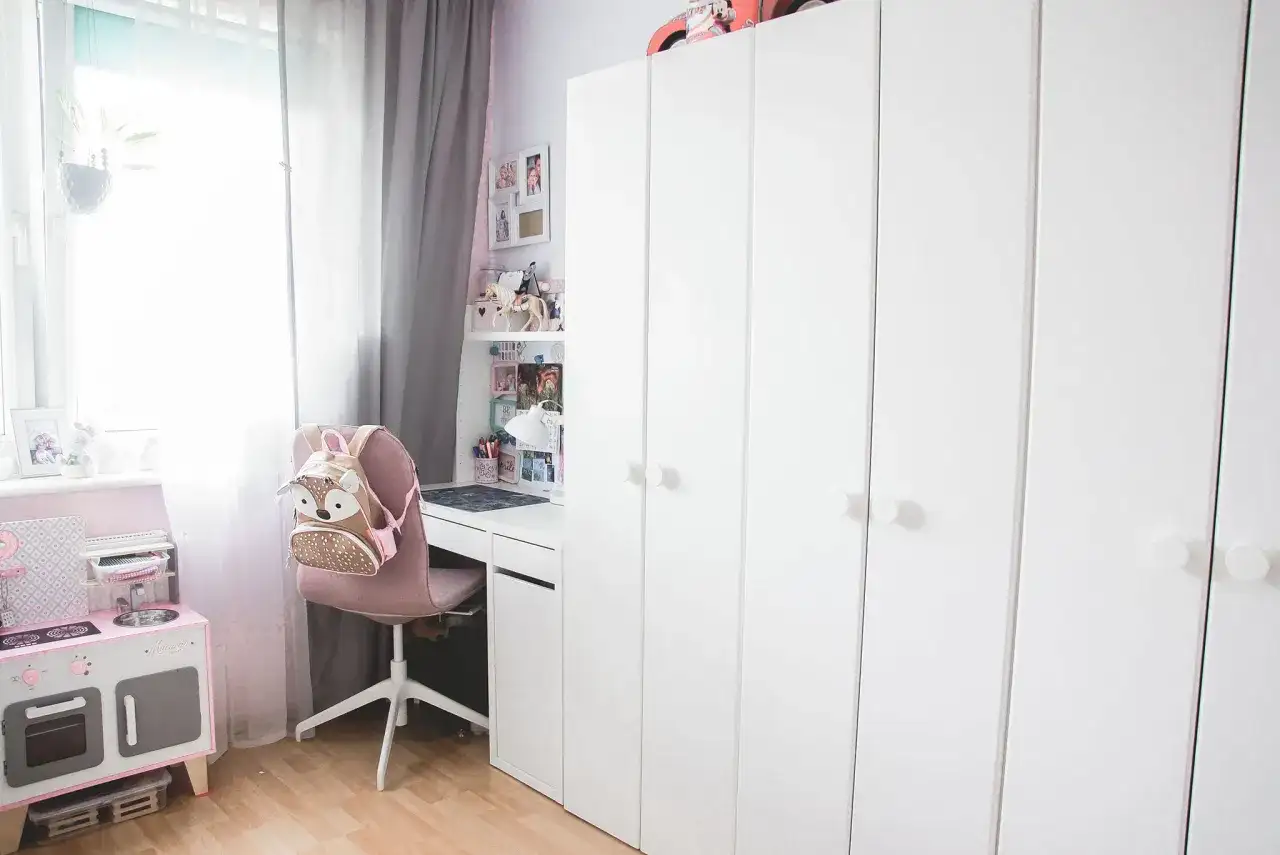 Urocza aranżacja małego pokoju z dużą białą szafą, biurkiem, różową kuchnią dla dzieci i plecakiem w kształcie sowy.