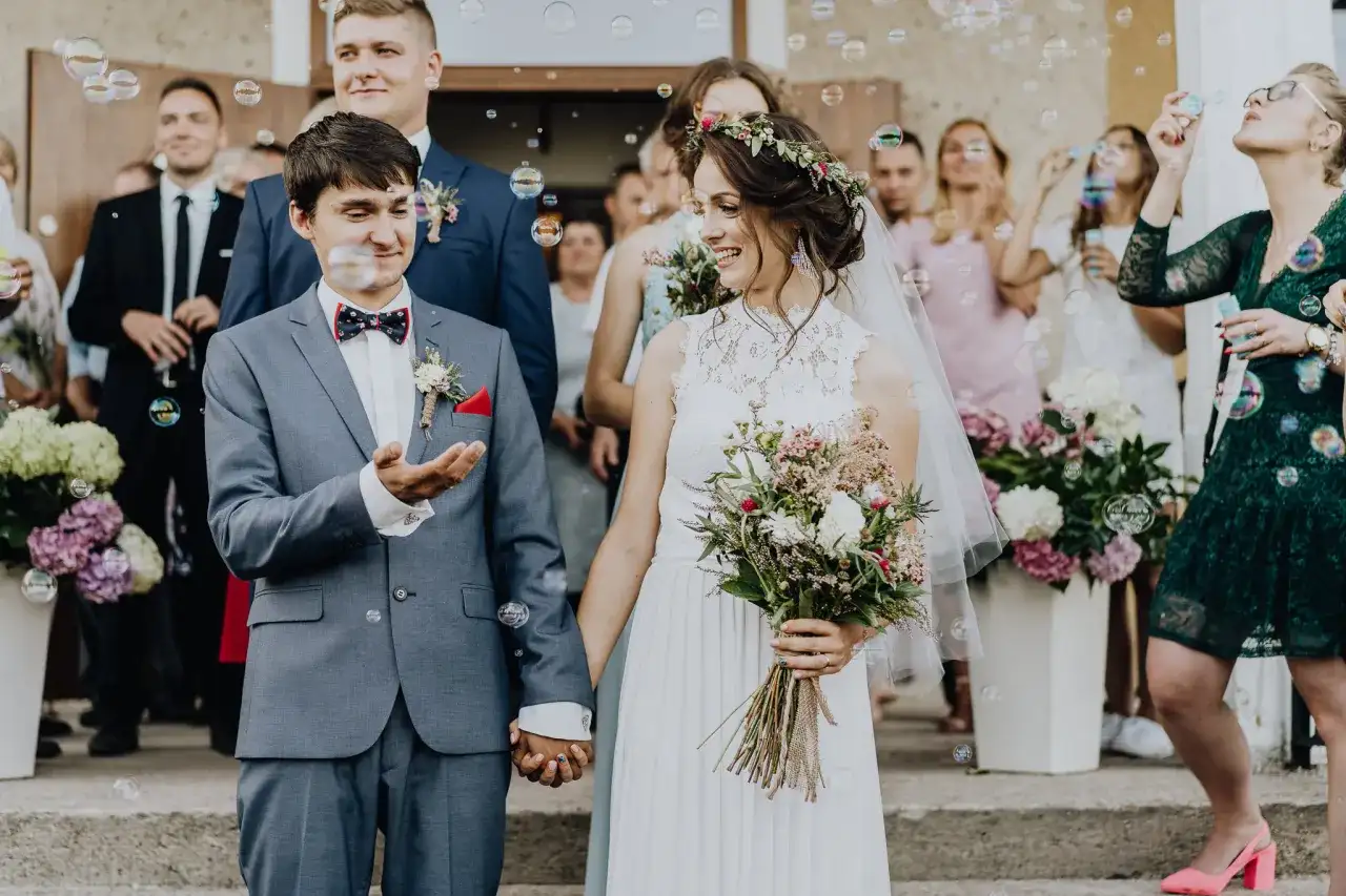 Para młoda trzymająca się za ręce, otoczona bańkami mydlanymi i gośćmi weselnymi.