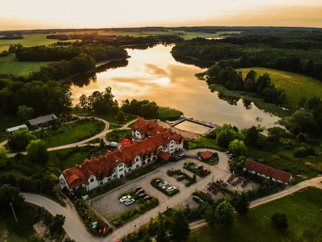 Malowniczy folwark mazurskie legendy nad jeziorem, z dachem krytym czerwoną dachówką, otoczony zielenią.