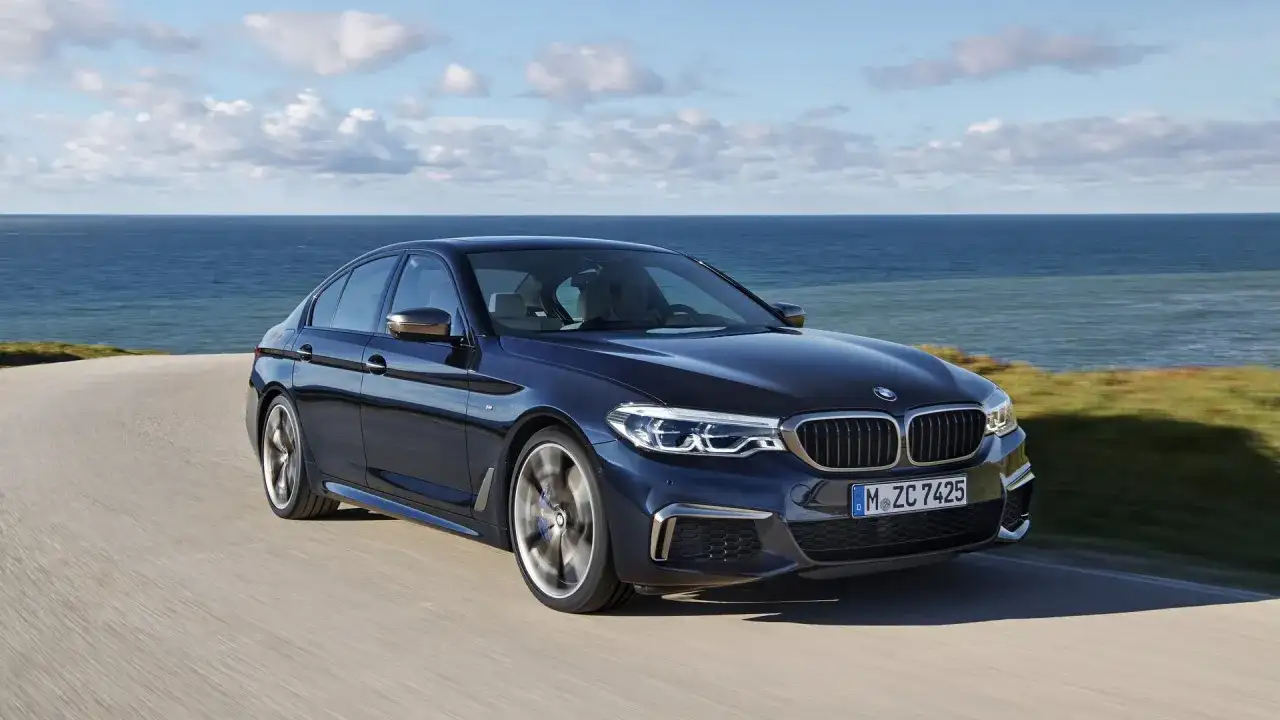 Ciemnoniebieskie BMW M550i sunie po krętej drodze nad brzegiem morza.