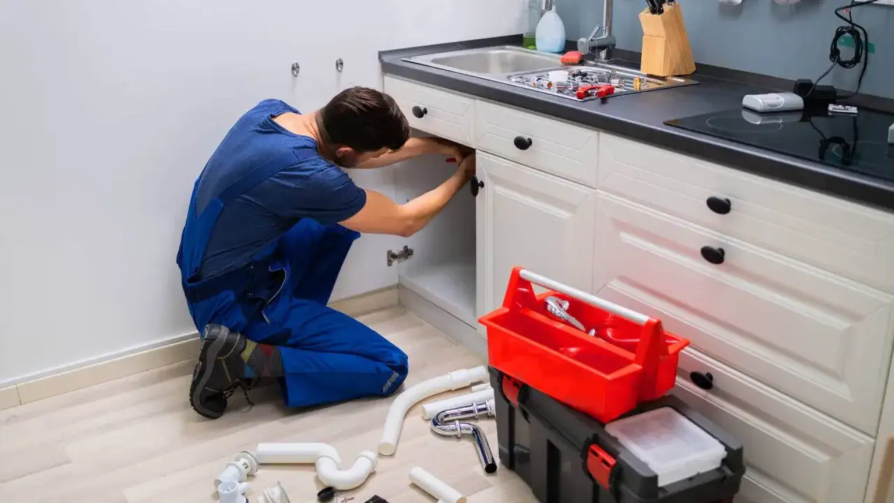 Hydraulik w niebieskim kombinezonie naprawia instalację pod zlewem w kuchni.