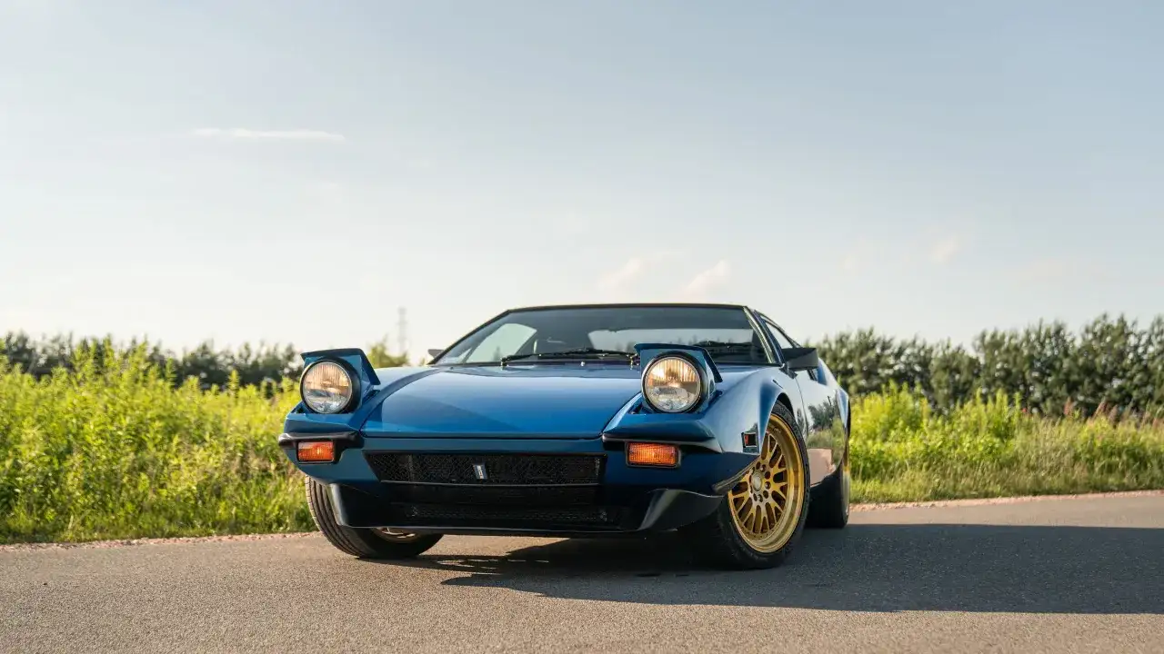 Niebieski De Tomaso Pantera z wysuwanymi światłami i złotymi felgami na tle zieleni i błękitnego nieba.