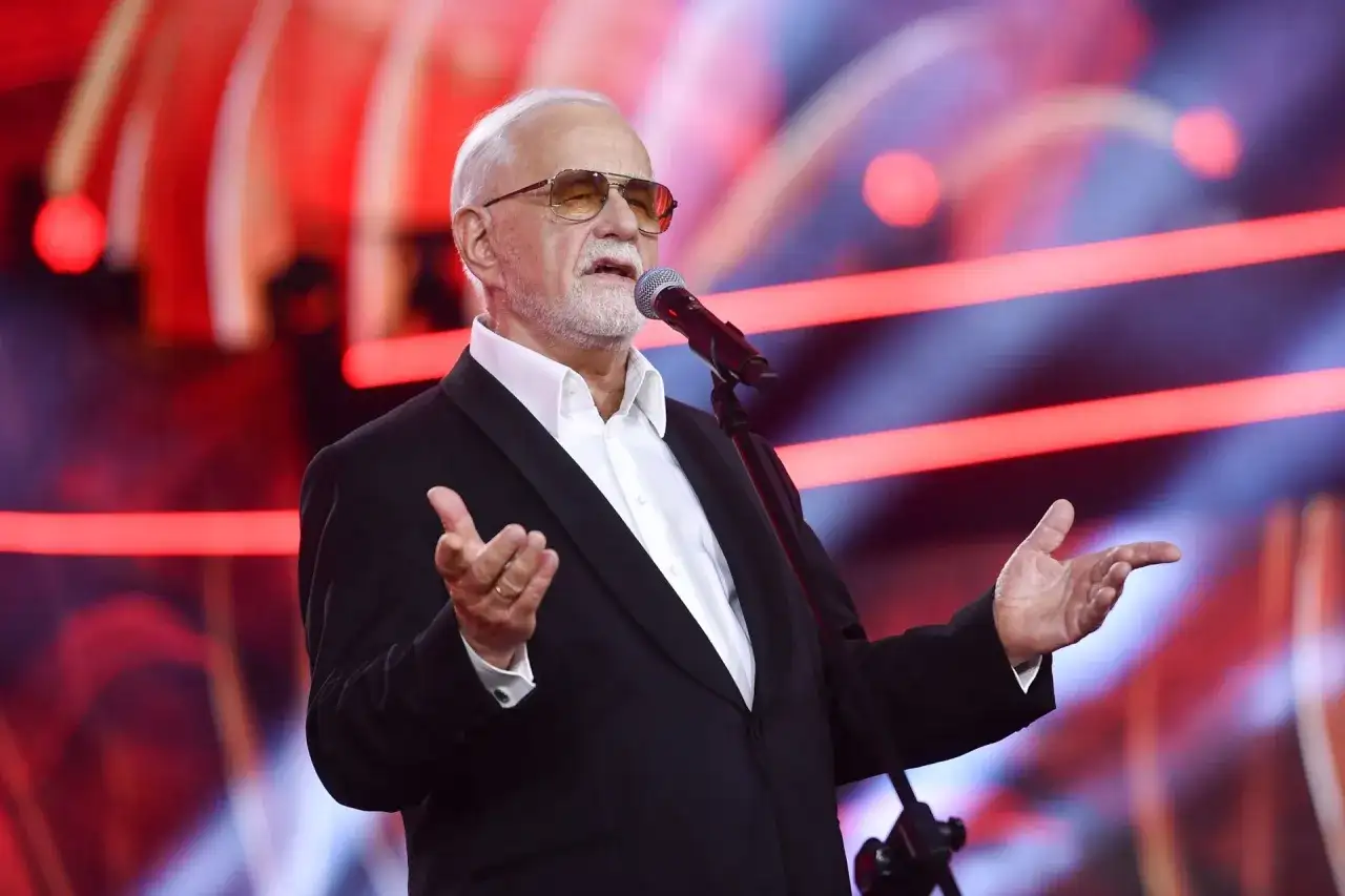 Jan Pietrzak w garniturze i okularach przeciwsłonecznych przemawia do mikrofonu na scenie.
