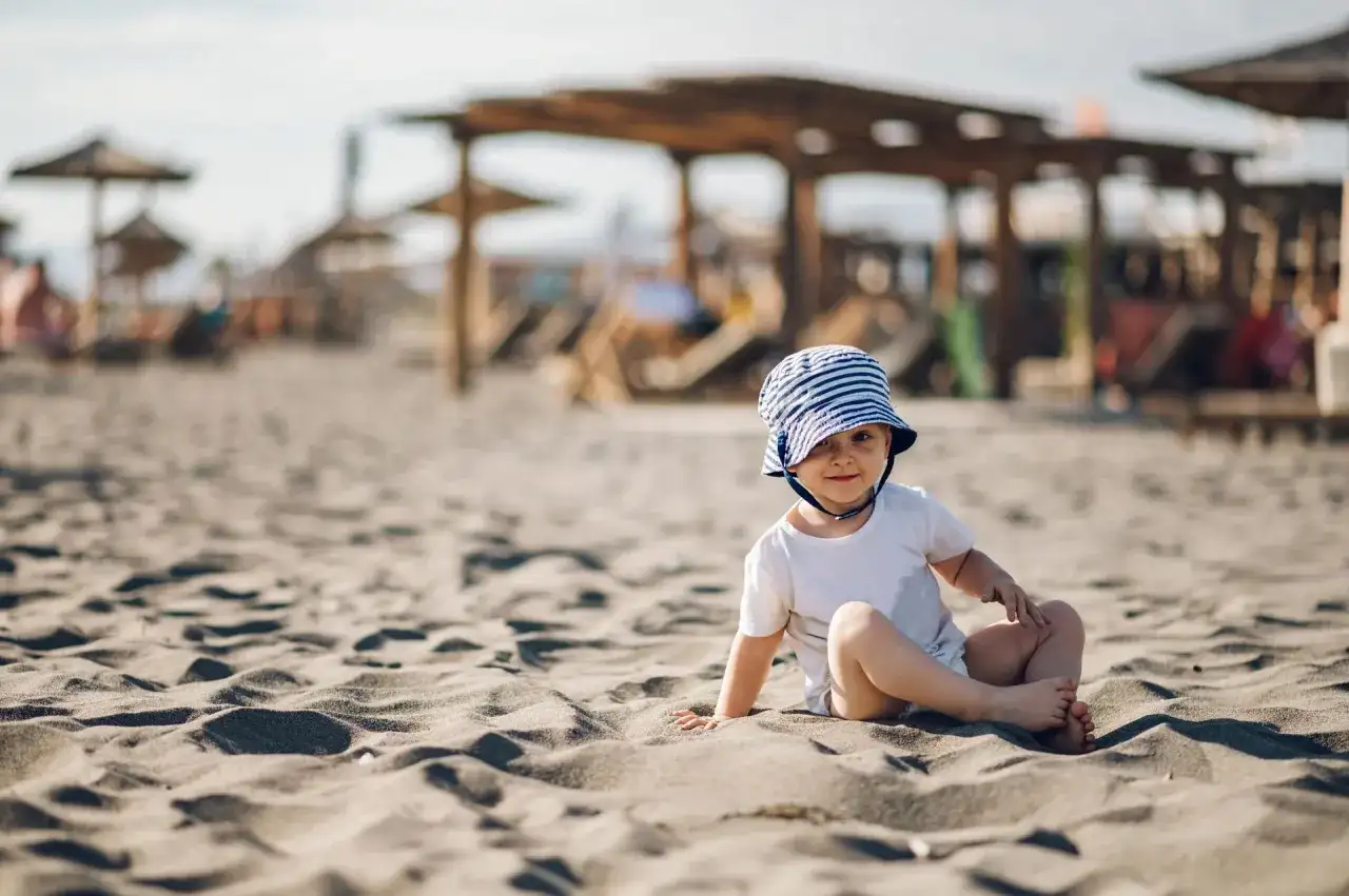 Uroczy maluch w pasiastym kapelusiku siedzi na piaszczystej plaży. Idealne miejsce, gdzie na wakacje z rocznym dzieckiem.