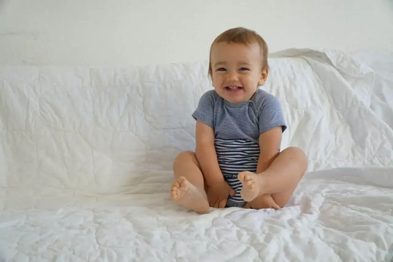 Lächelndes Baby im gestreiften Body sitzt auf weißer Decke.