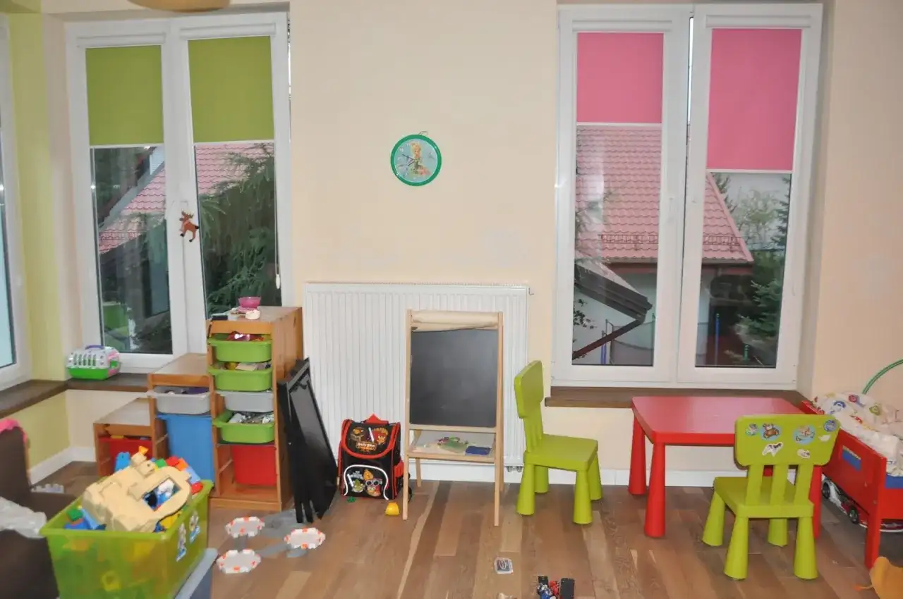 Pokój dziecięcy z zabawkami, stolikiem i krzesełkami. Okna z zielonymi i różowymi roletami, a za nimi widać dachy domów. Trofast inspiracje dla małych artystów.