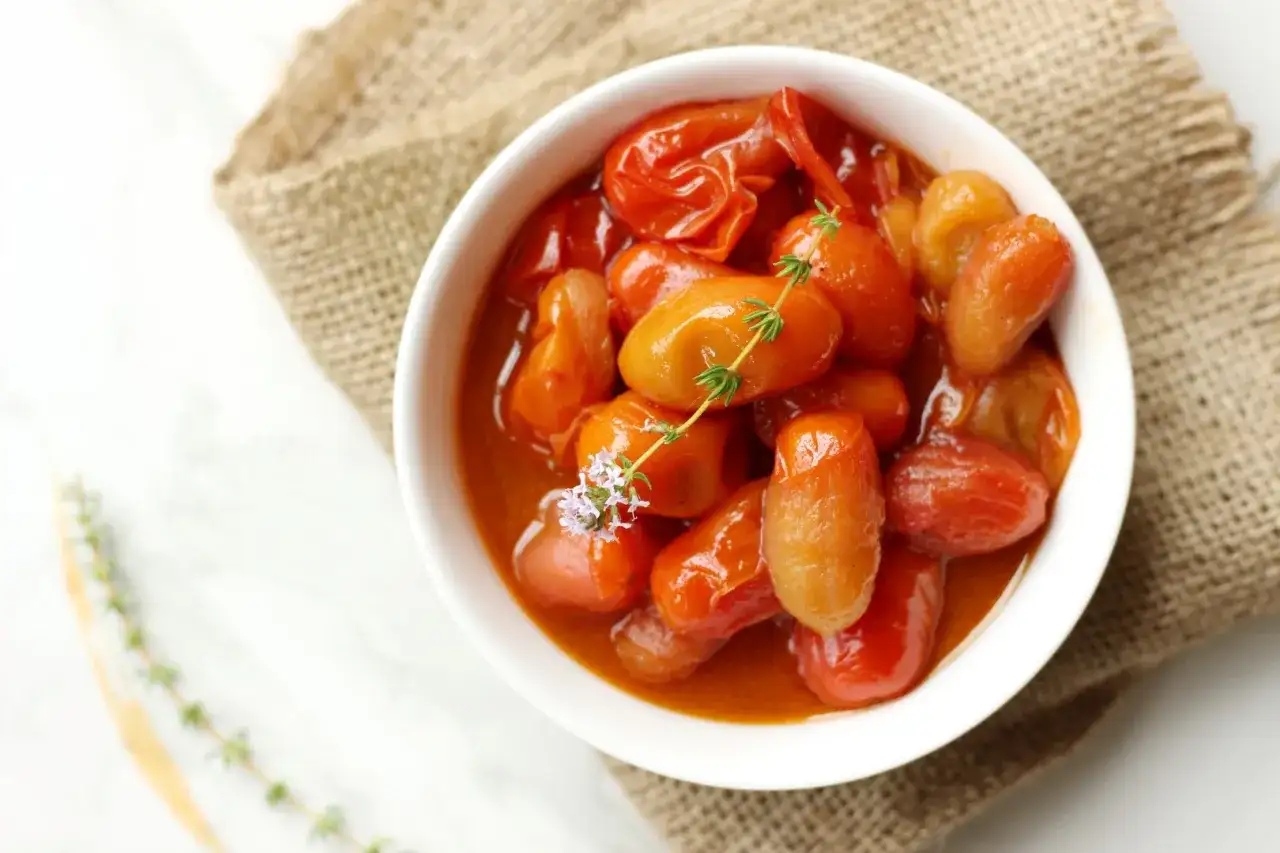 Un tazón blanco lleno de tomates cherry confitados, con una ramita de tomillo y una flor morada.