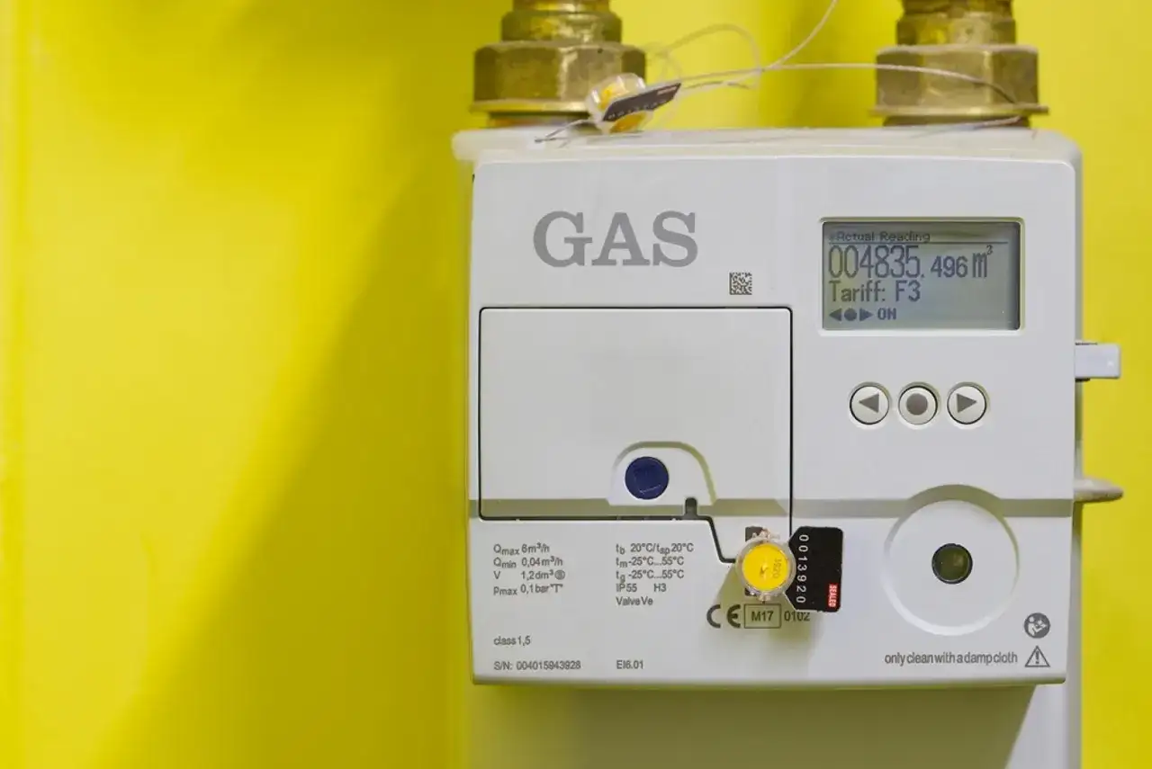 Medidor de gas blanco con pantalla digital y conexiones doradas sobre fondo amarillo.
