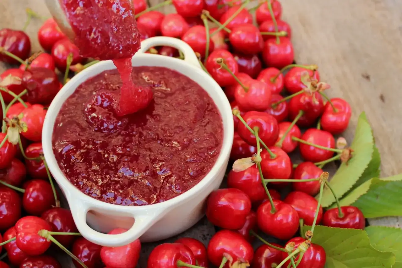 Cuchara vierte mermelada de cerezas casera en un cuenco blanco, rodeado de cerezas frescas y hojas verdes.