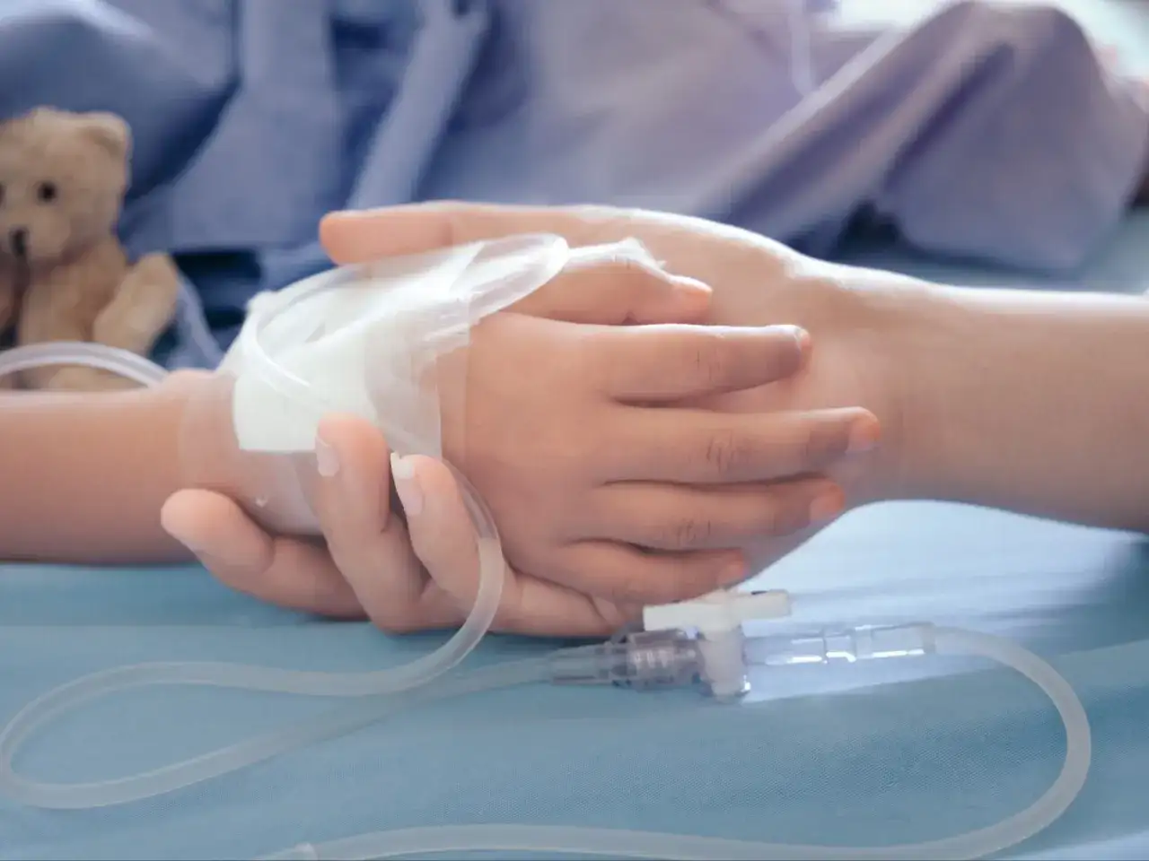 Dłoń dorosłego obejmuje dłoń dziecka z wenflonem. W tle pluszowy miś. To obraz wsparcia, gdy potrzebny jest zasilek pielegnacyjny.