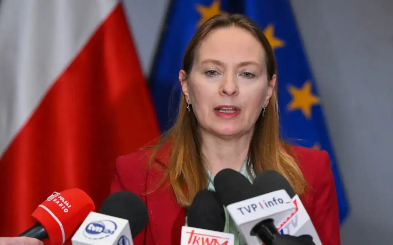 Kobieta w czerwonej marynarce przemawia do mikrofonów. W tle flagi Polski i UE. To po a pis porównanie.