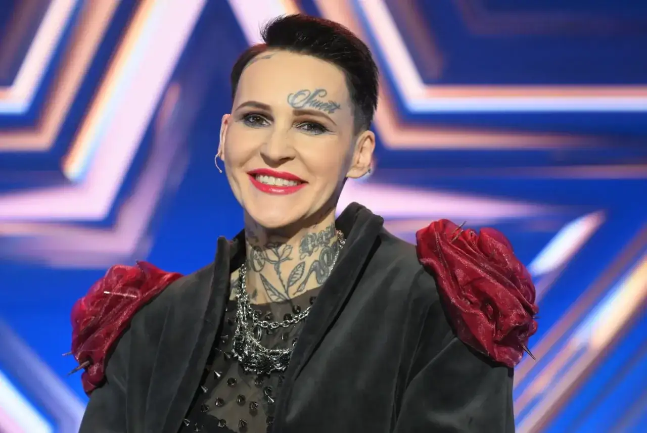 Agnieszka Chylińska z tatuażami na twarzy i szyi, w czarnej marynarce z czerwonymi ozdobami na ramionach.