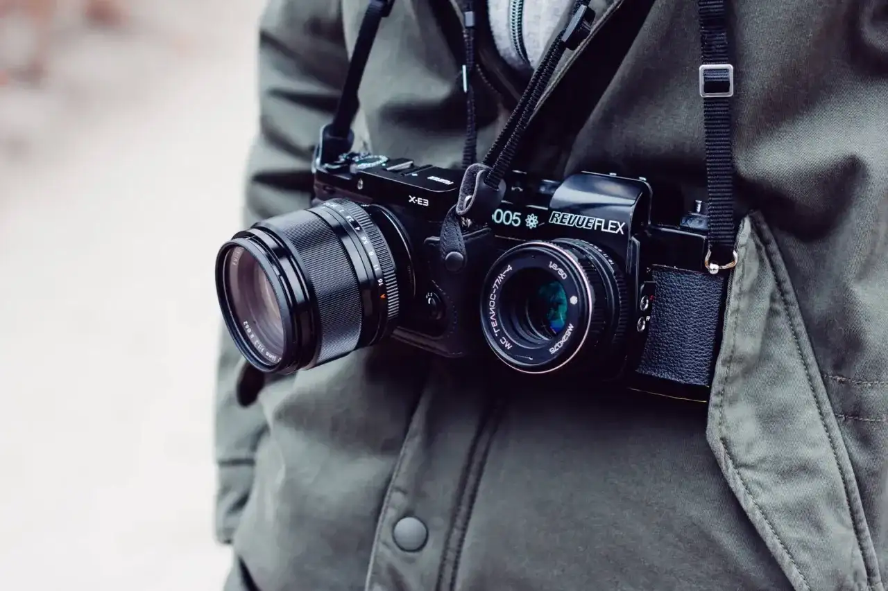 Osoba w zielonej kurtce nosi na szyi dwa aparaty fotograficzne: jeden cyfrowy, drugi analogowy.