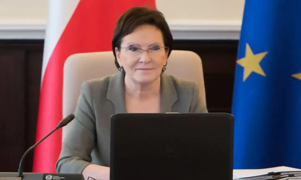 Ewa Kopacz, premier, siedzi przy stole z laptopem, obok flagi Polski i Unii Europejskiej.