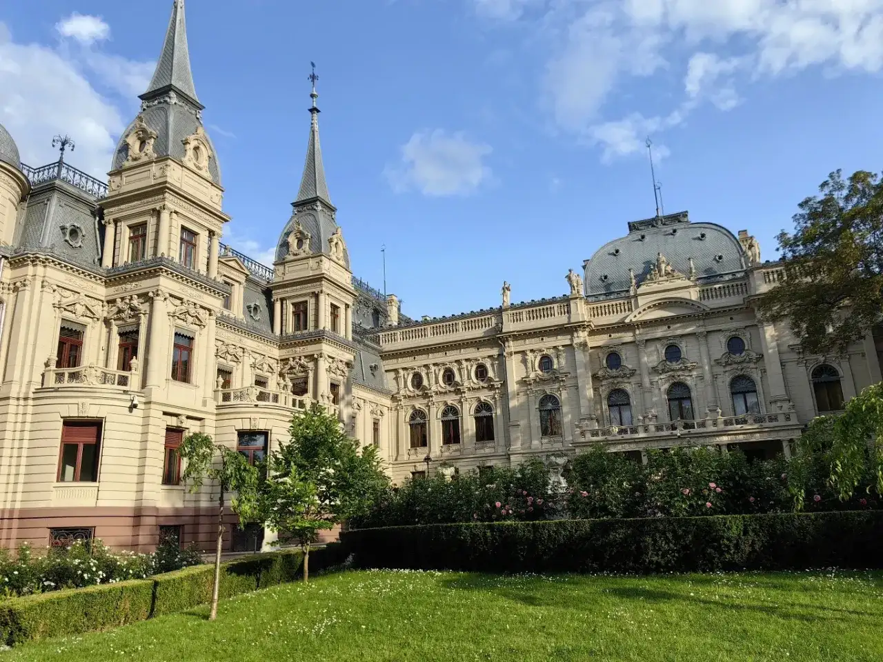 Imponujący pałac poznańskiego, z wieżyczkami i bogatymi zdobieniami, otoczony zielonym ogrodem z kwitnącymi krzewami róż.