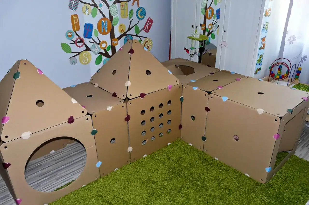 Zabawki z kartonu DIY tworzą domek z otworami i okrągłym wejściem. Kolorowe serduszka zdobią kartonowe ściany.