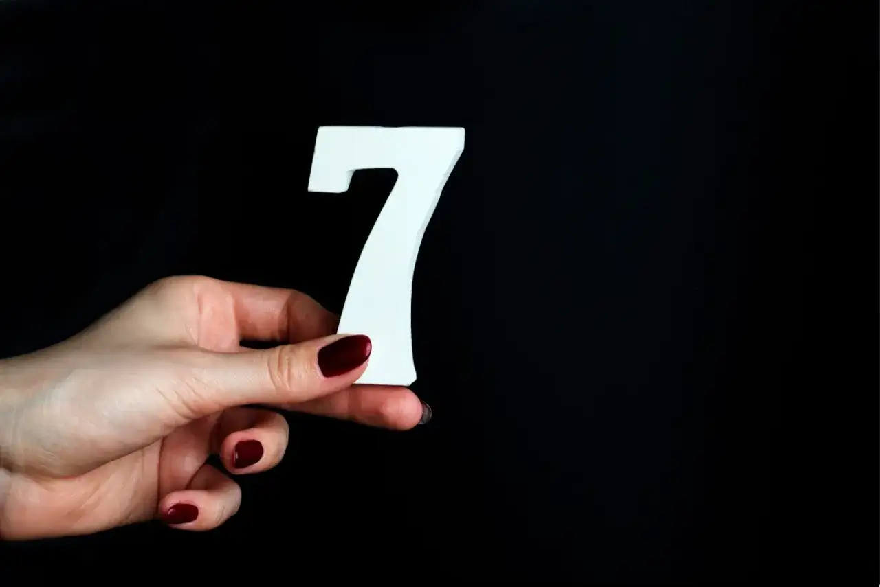 Dłoń z ciemnoczerwonym lakierem trzyma białą cyfrę 7, symbolizującą numerologiczną 7.