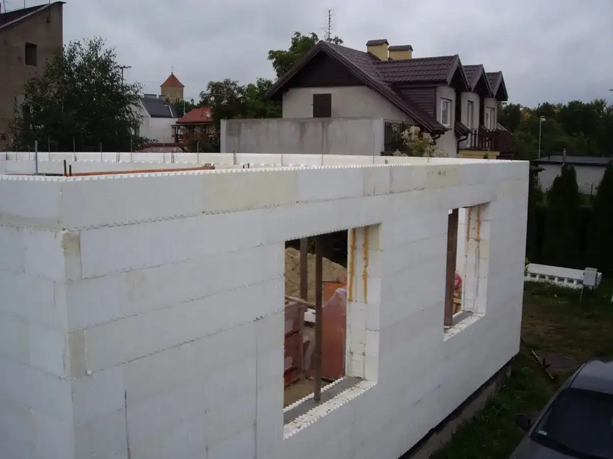 Budowa domu ze styropianu. Białe bloki tworzą ściany z otworami okiennymi. W tle widać inne budynki i drzewa.