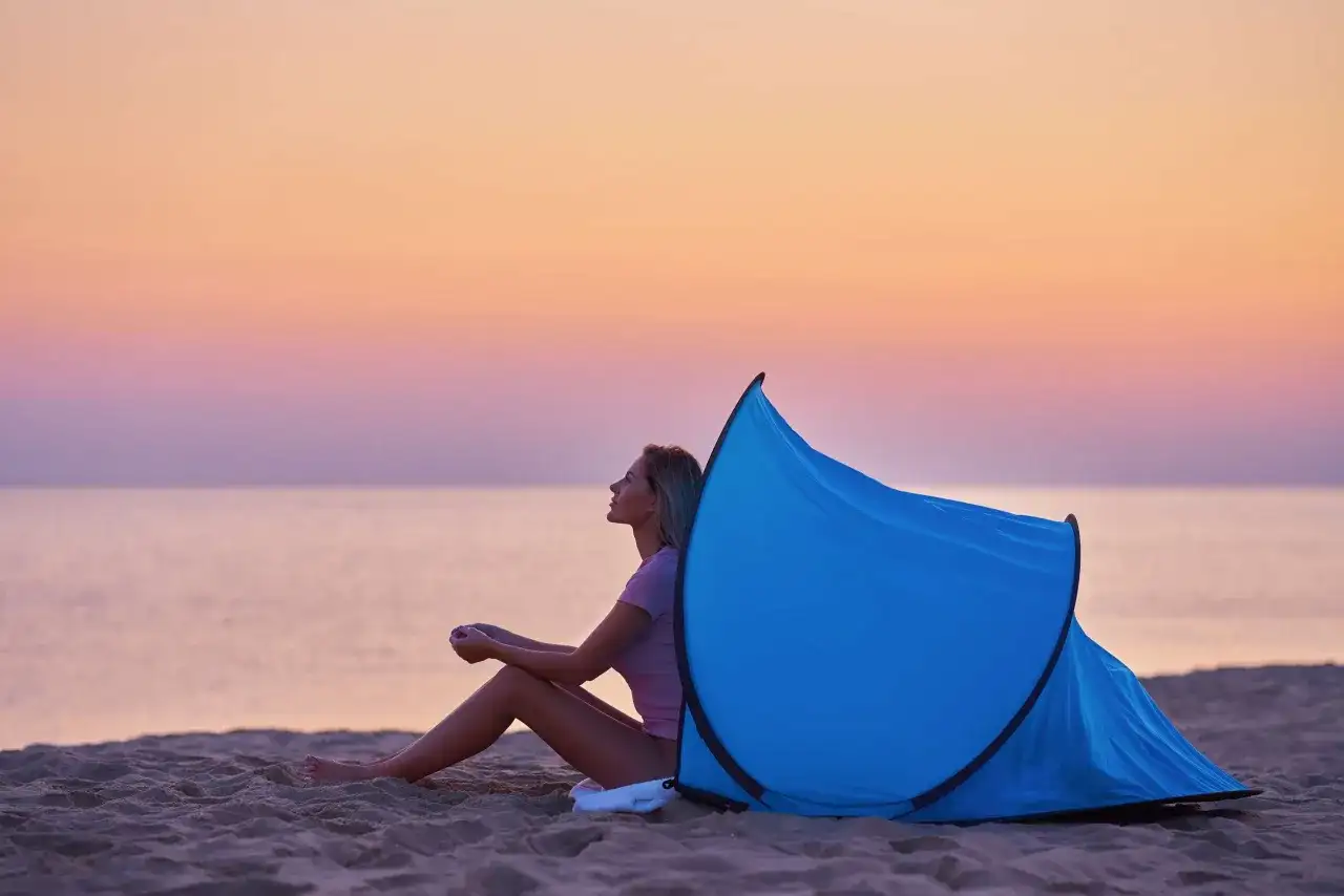 Kobieta siedzi na plaży przy rozłożonym namiocie plażowym. Zachód słońca nad morzem. Jak złożyć namiot plażowy instrukcja.