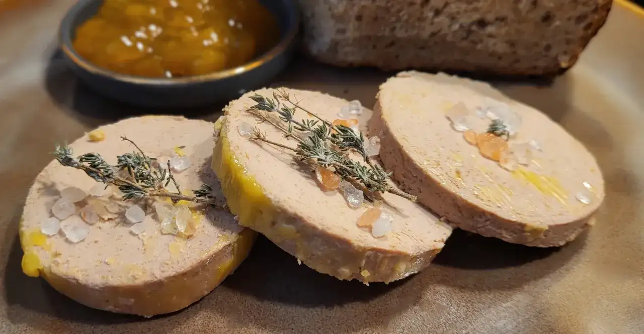 Trzy plastry foie gras, posypane solą i gałązkami tymianku, obok dżemu i chleba. Foie gras co to? Delikatesowy pasztet z gęsiej lub kaczej wątróbki.