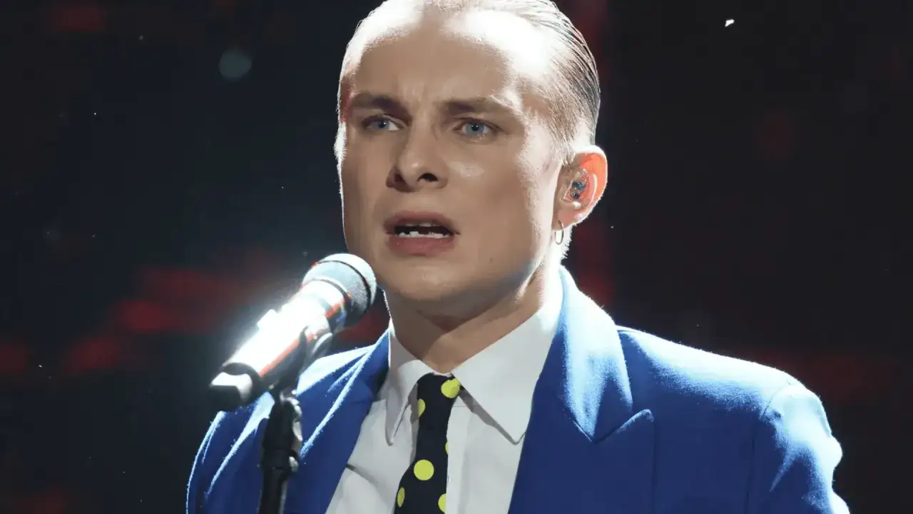 Ralph Kaminski, w niebieskiej marynarce i krawacie w grochy, śpiewa do mikrofonu. Jego partner, wokalista, jest w centrum uwagi.