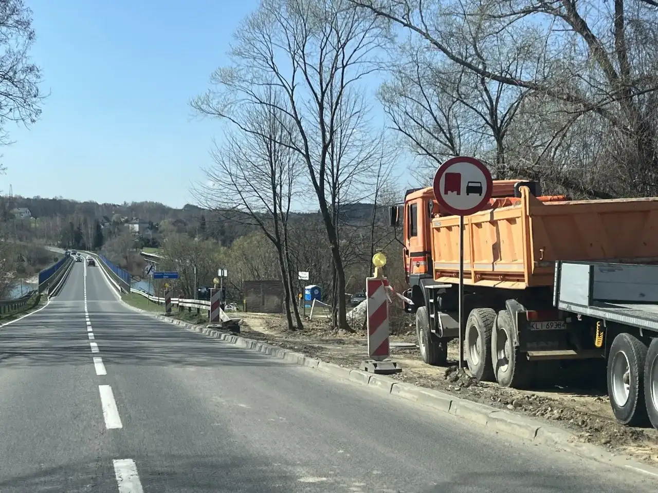Pomarańczowa wywrotka MAN stoi przy drodze, gdzie trwa remont. Znak zakazu wyprzedzania dla pojazdów ciężarowych. Velo Raba w tle.