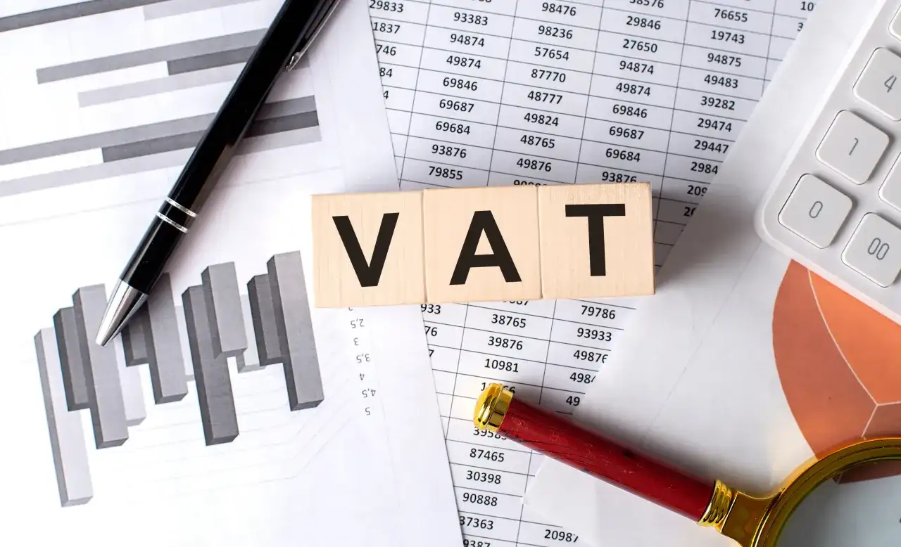 Drewniane klocki z napisem VAT leżą na dokumentach z wykresami i tabelami. Jak odliczyć VAT z faktury? Pióro i lupa sugerują analizę.