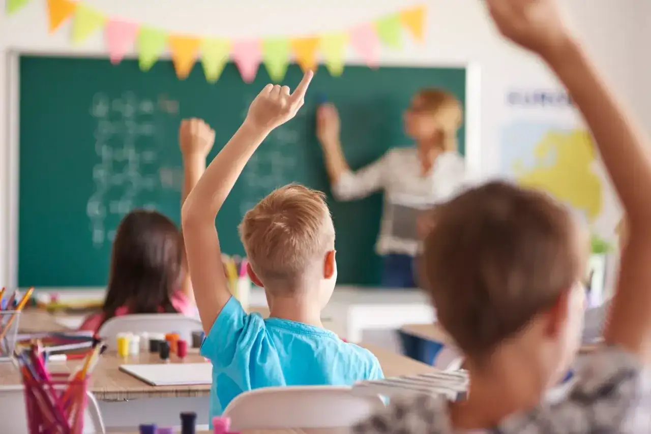 Uczniowie z podniesionymi rękami słuchają uważnie, co mówi wychowawca przy tablicy.