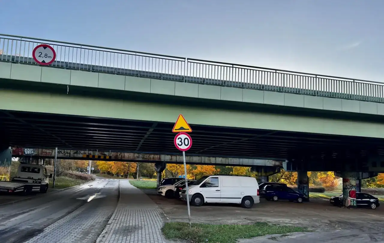 Pod estakadą zaparkowane są samochody. Widoczny znak ograniczenia wysokości 2,8m i znak ograniczenia prędkości do 30 km/h.