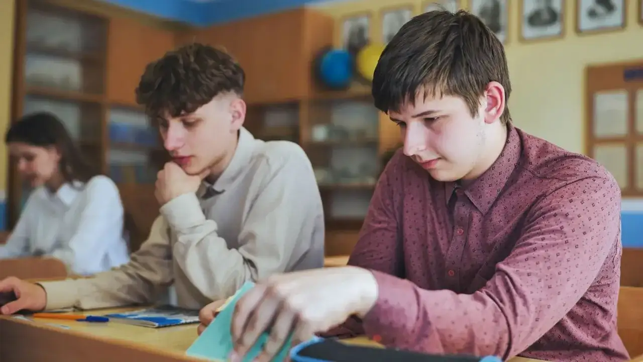 Uczniowie siedzą przy ławkach w klasie, jeden z nich przegląda notatki.