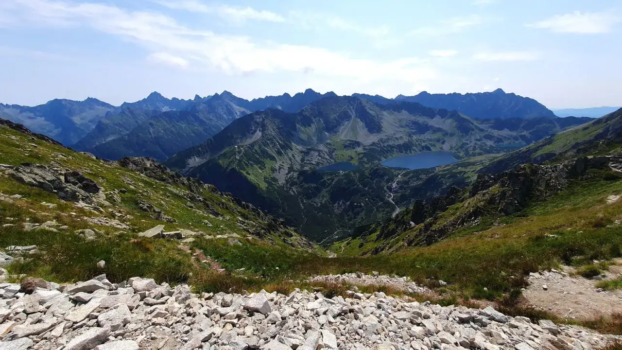 Widok na Tatry z kamienistego zbocza. W dole jeziora i kręte szlaki na krzyżne, otoczone zielenią i skalistymi szczytami.
