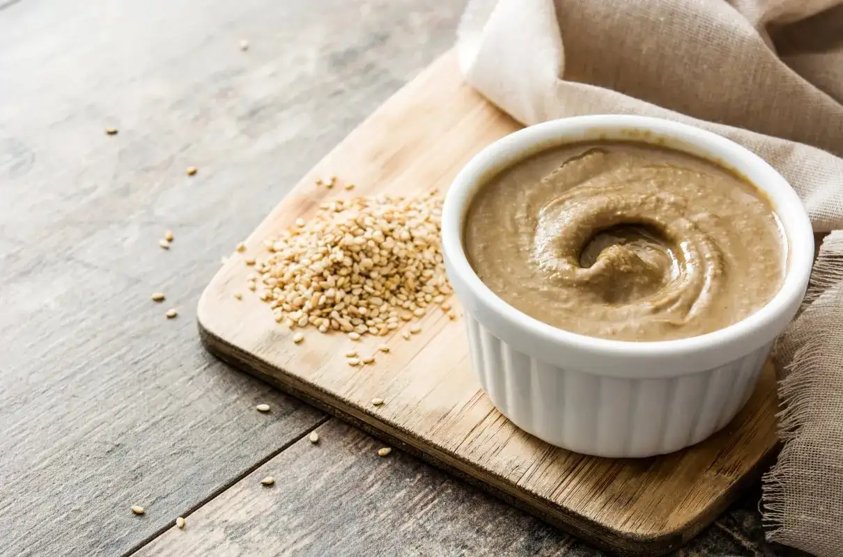 Domowe tahini jak zrobić? Ziarna sezamu i gotowa pasta na desce.
