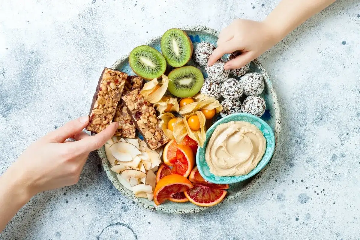 Talerz przekąsek na imprezę: batoniki, kiwi, kulki kokosowe, pomarańcze, physalis i pasta. Dzieci sięgają po smakołyki.