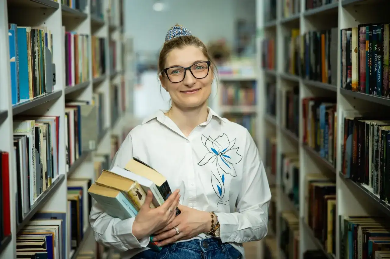 Bibliotekarka z okularami trzyma stos książek. W tle regały pełne literatury. Czy wakat co to znaczy?