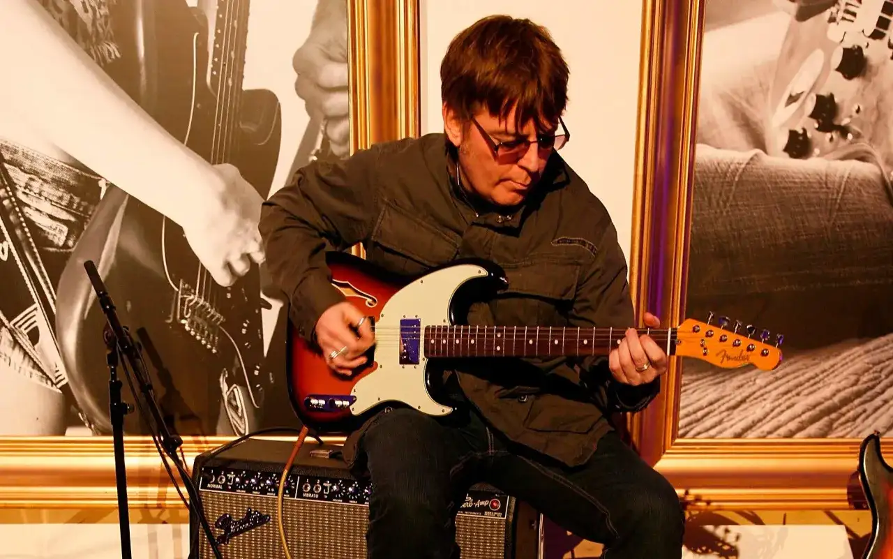 Mężczyzna w okularach gra na gitarze elektrycznej Fender, siedząc na wzmacniaczu.