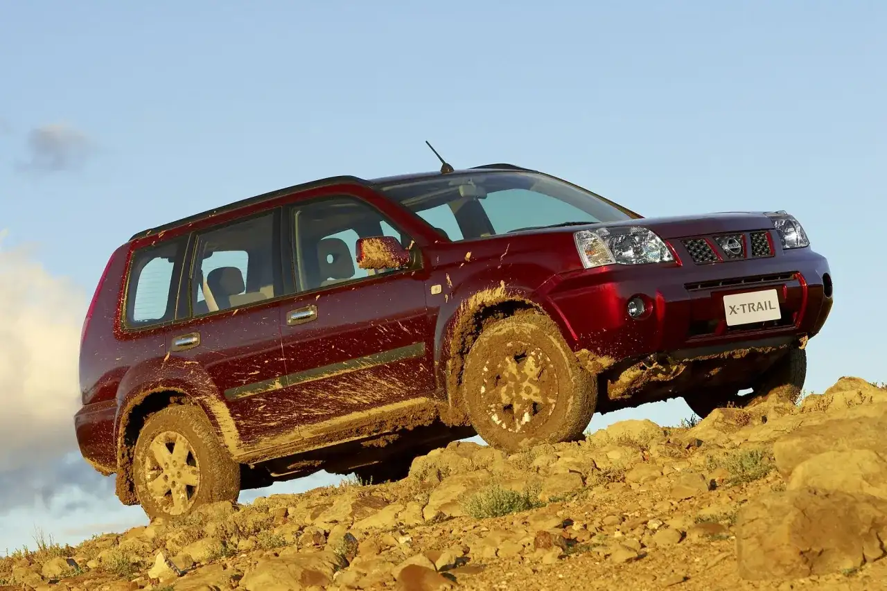 Czerwony Nissan X-Trail ubrudzony błotem podjeżdża pod skaliste wzniesienie.
